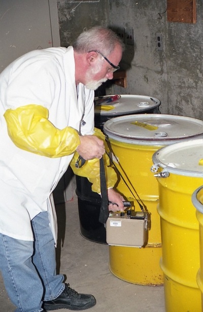 Chuck Surre, a University of Rochester radiation safety technician is shown here performing a radiation survey around some drums of just-compacted radioactive waste in the waste storage room.